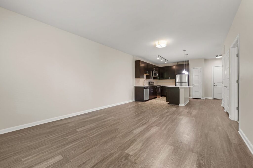 Open-concept kitchen with dark wood cabinets and a peninsula, overlooking an empty living area with light-colored walls and wood-look flooring.