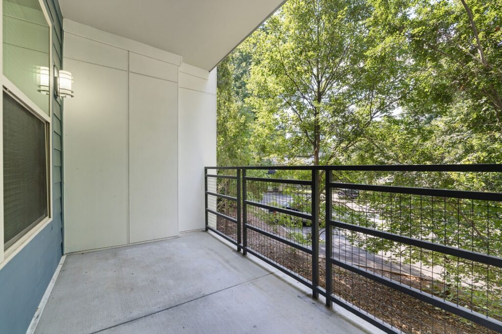 A covered apartment balcony with concrete flooring and a black metal railing overlooking trees.